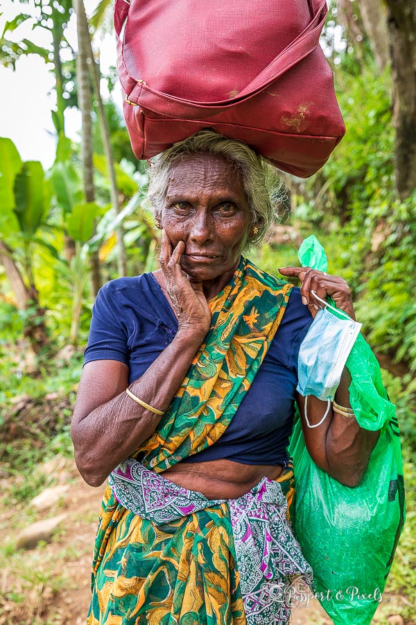 An elderly woman in a village in the Knuckles Mountain Range. She's wearing a blue t-shirt and balancing a red bag on her head.