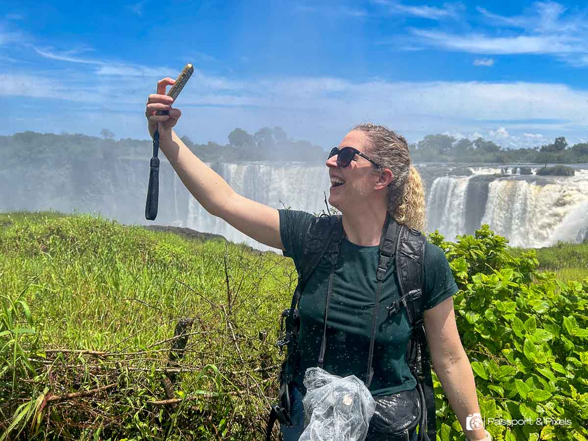 The author, me, wet from spray from the waterfall, standing in front of Victoria Falls, holding up my phone to take a selfie