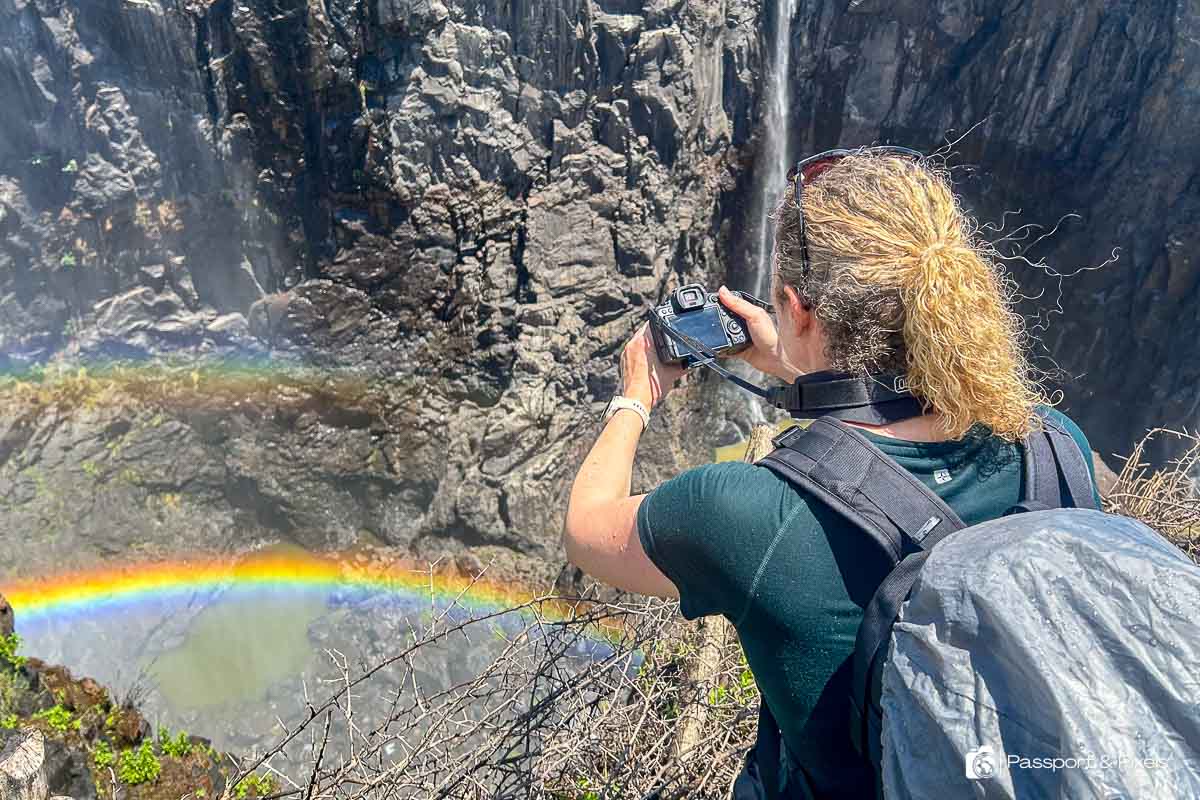 Me, the author, with my back to the camera, looking out over Victoria Falls waterfall, and holding out my camera to photograph a brightly coloured rainbow