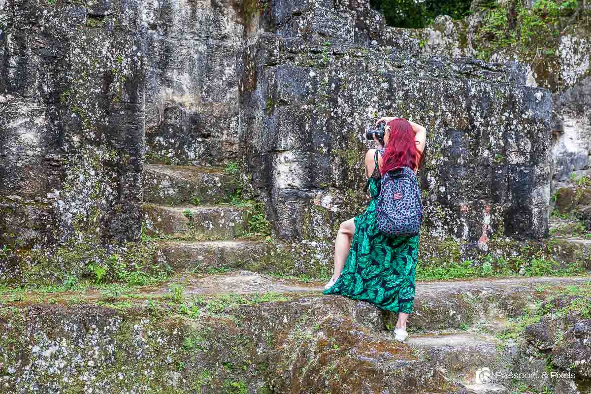 A woman in a green dress with red hair taking a photo of some ruins at Tikal in Guatemala